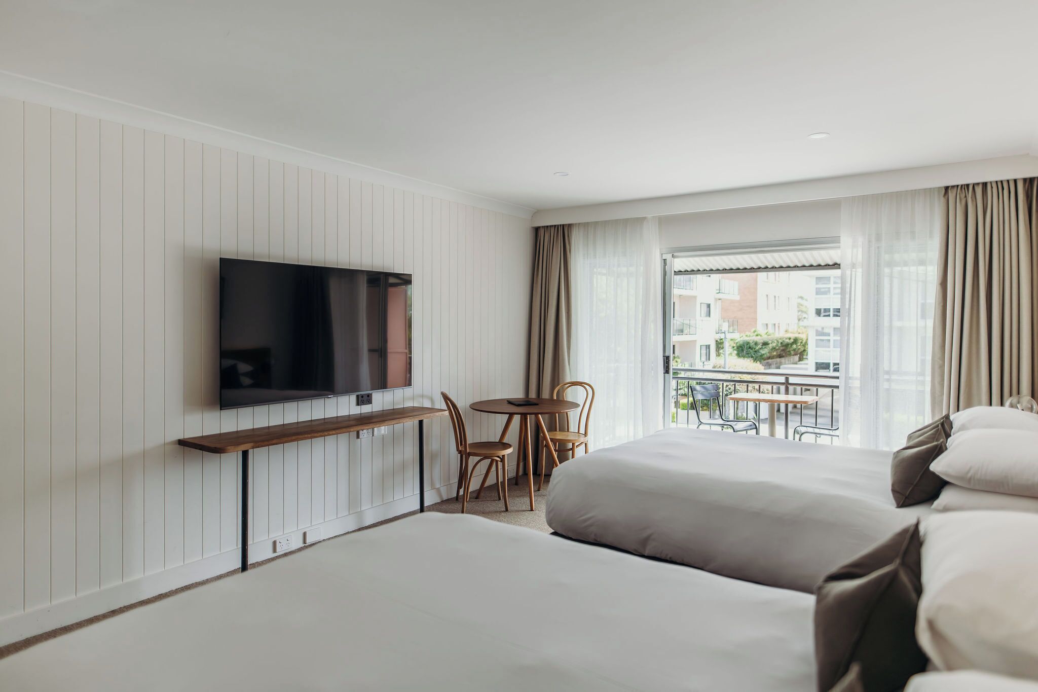 Une chambre d’hôtel avec deux lits, un téléviseur fixé au mur, une petite table avec des chaises et un balcon donnant sur des immeubles.
