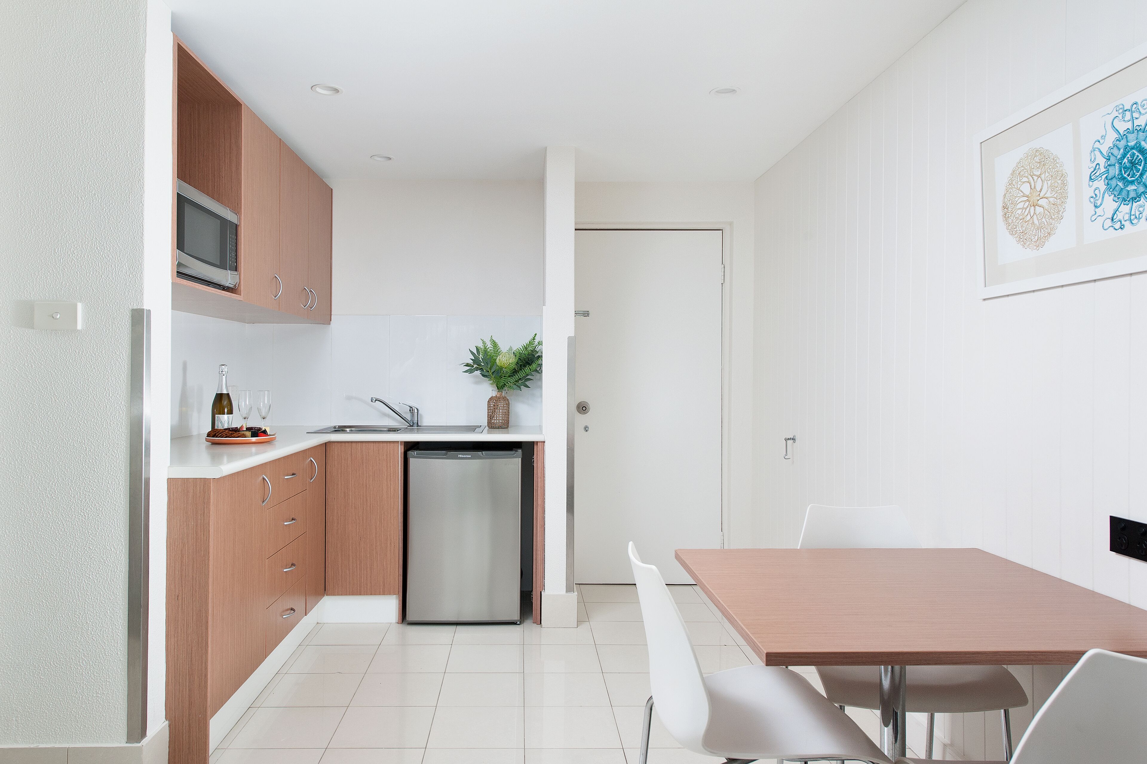 A compact kitchen with wooden cabinets, a built-in microwave, and a dishwasher. There is a dining area with a wooden table and white chairs.