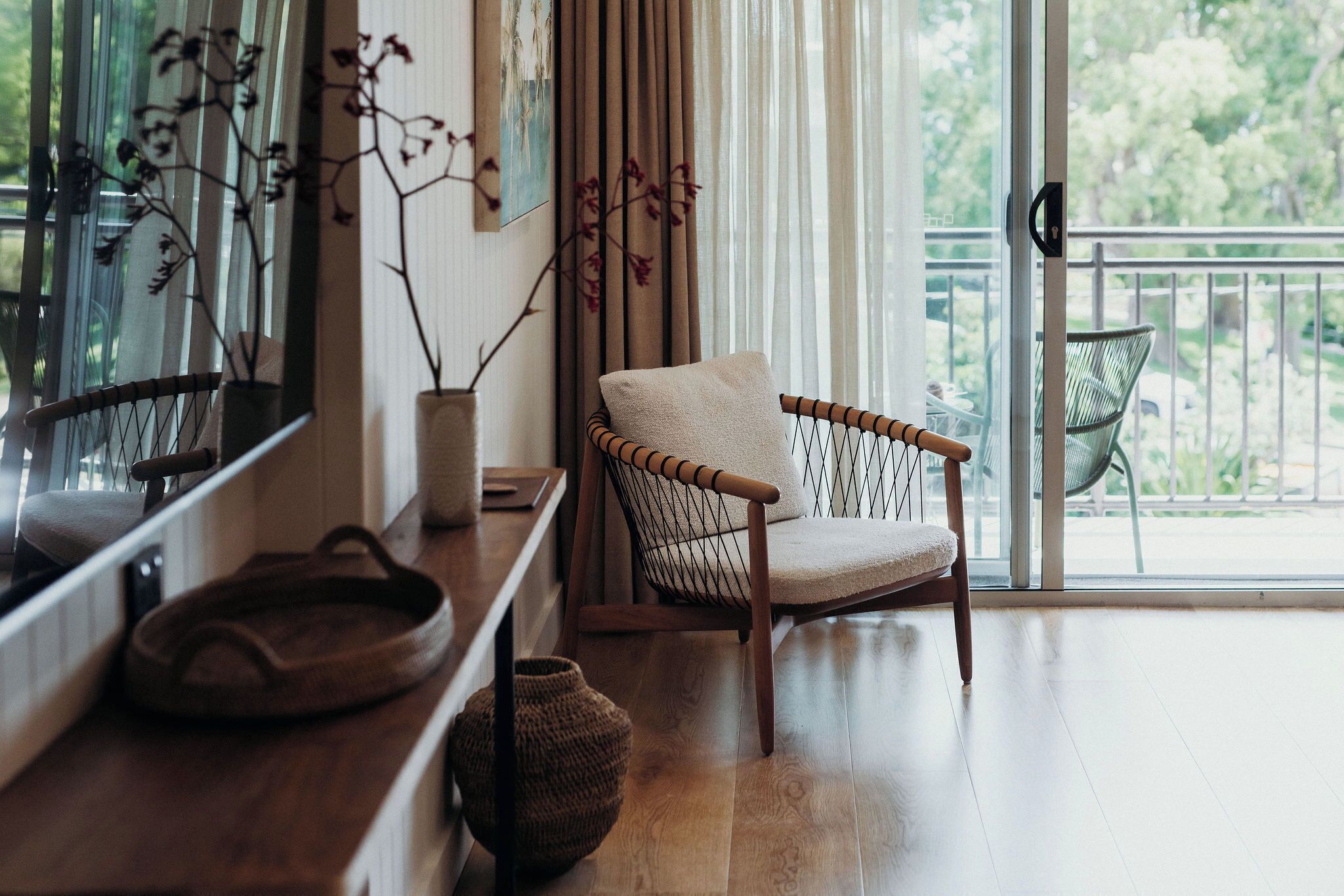 Une pièce avec un plancher en bois, une chaise en osier avec un coussin, un panier tressé et une grande fenêtre avec des rideaux.