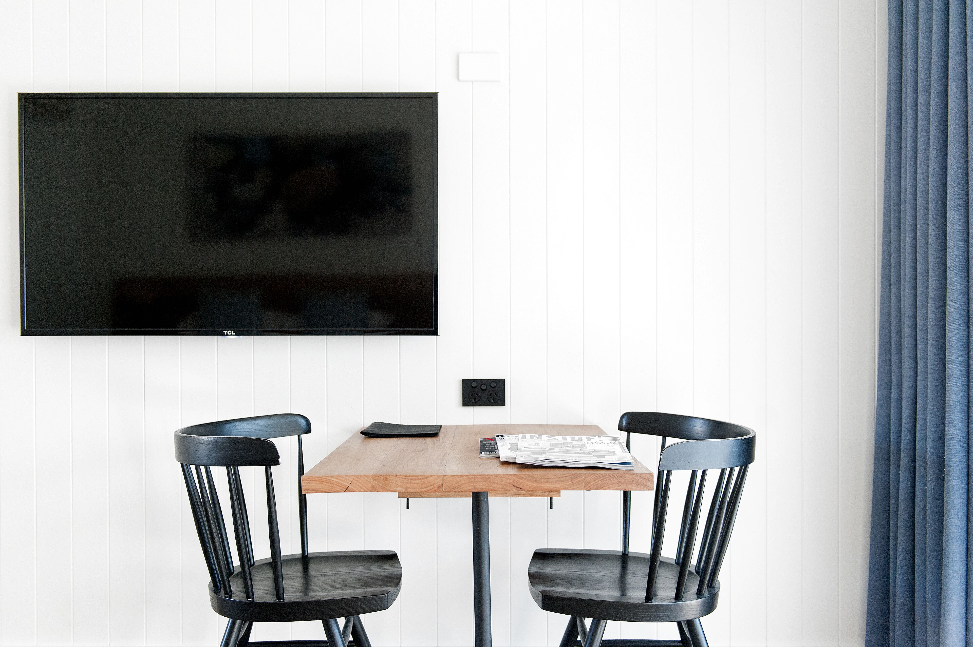 A dining area with a wooden table, two black chairs, a flat-screen TV mounted on the wall, and a power outlet below it.