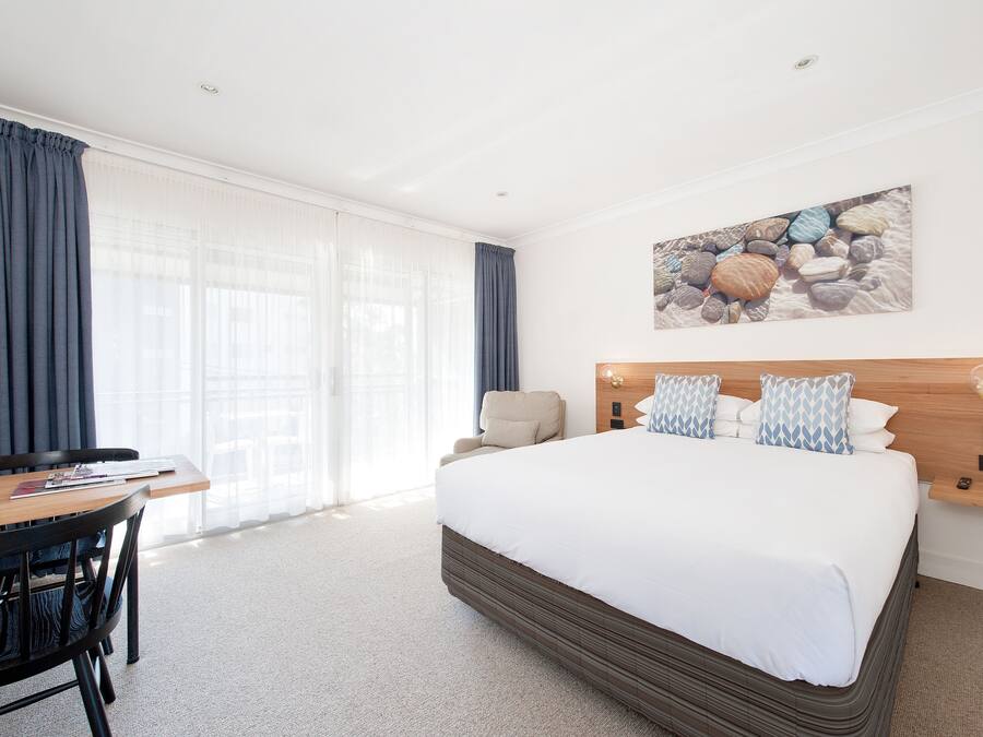 A hotel room with a large bed, a wooden desk, a chair, and a view of the outdoors through sheer curtains.