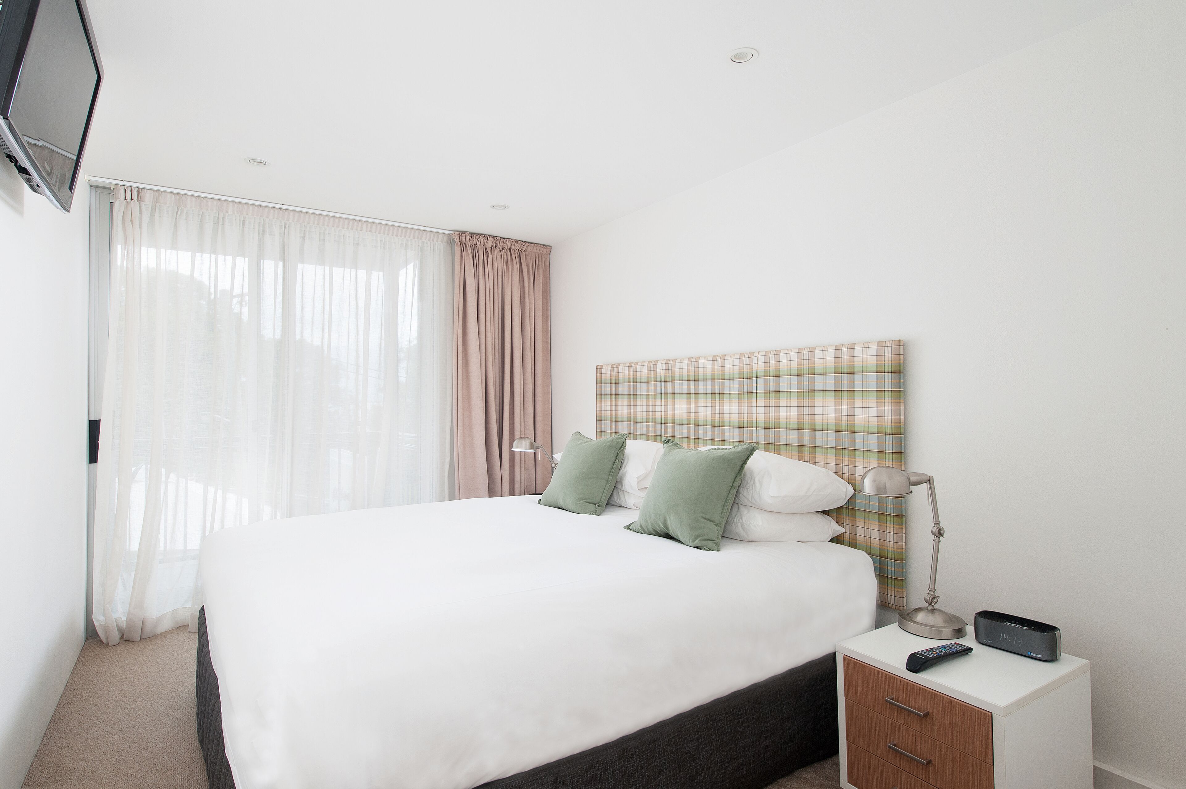 A neatly made bed with white linens and green pillows, a bedside table with a lamp and remote control, and a plaid-patterned headboard.