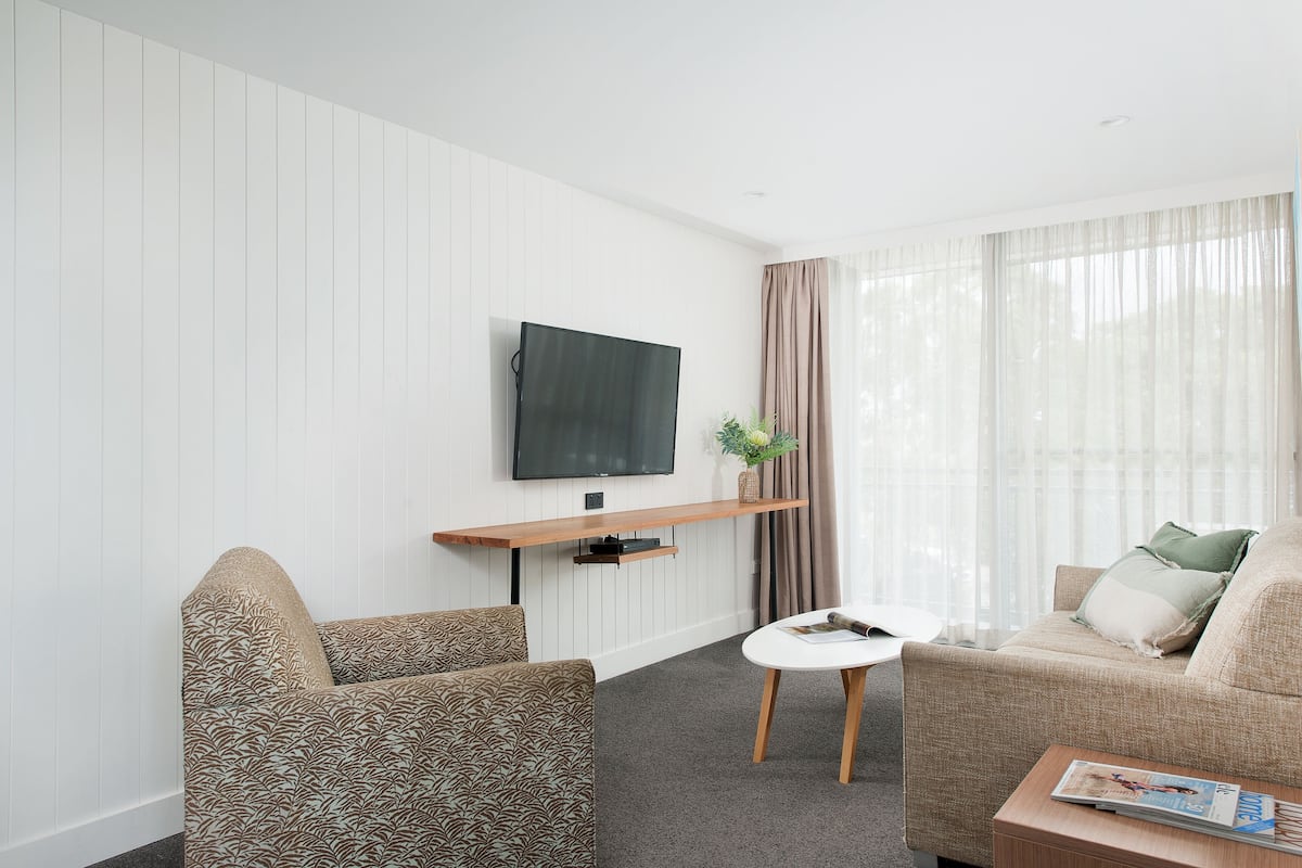 En moderne stue med et fladskærms-tv monteret på væggen, en træhylde nedenunder og en beige sofa med mønstrede puder.