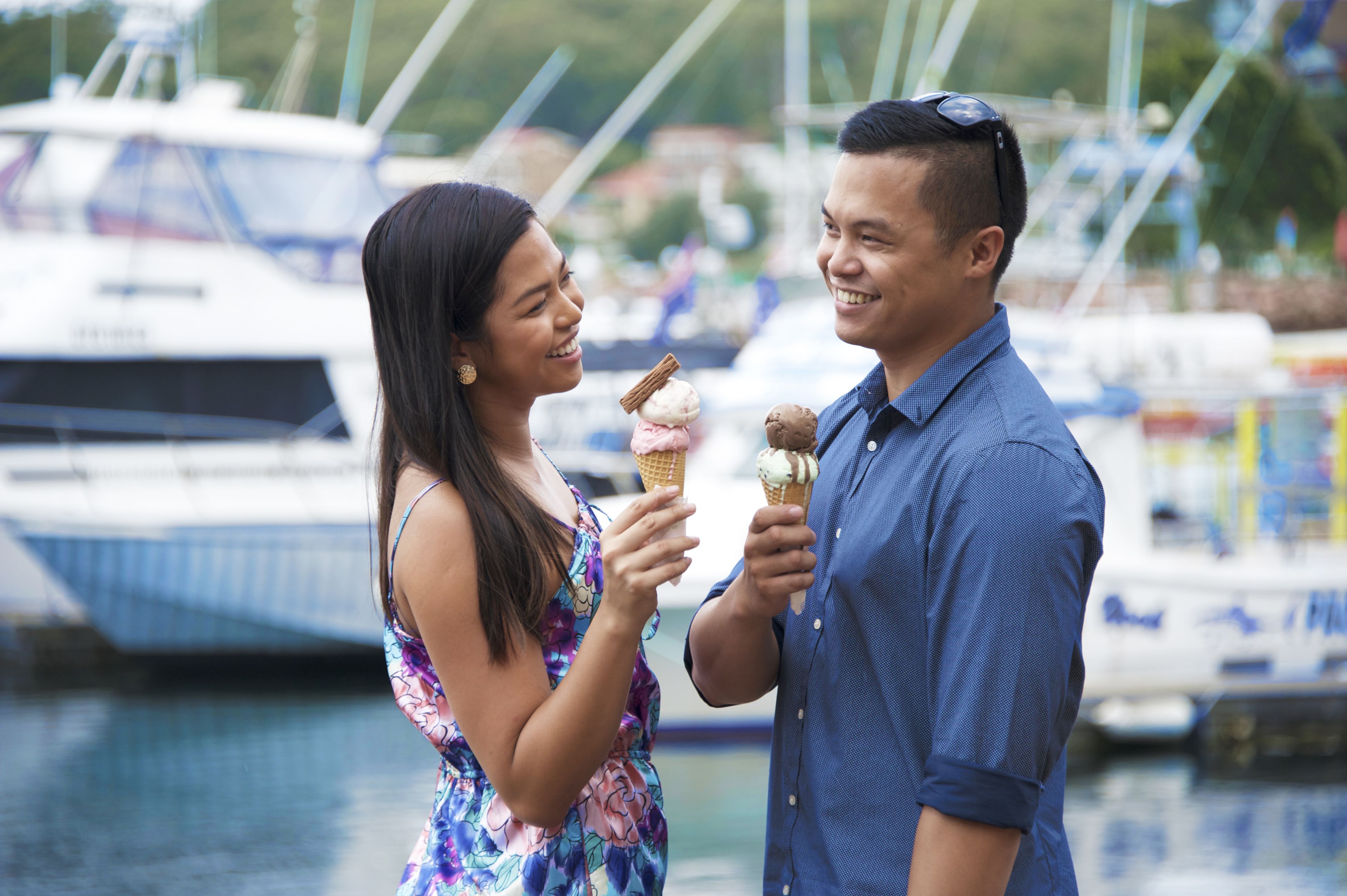 Two people holding ice cream cones by a marina with boats in the background.