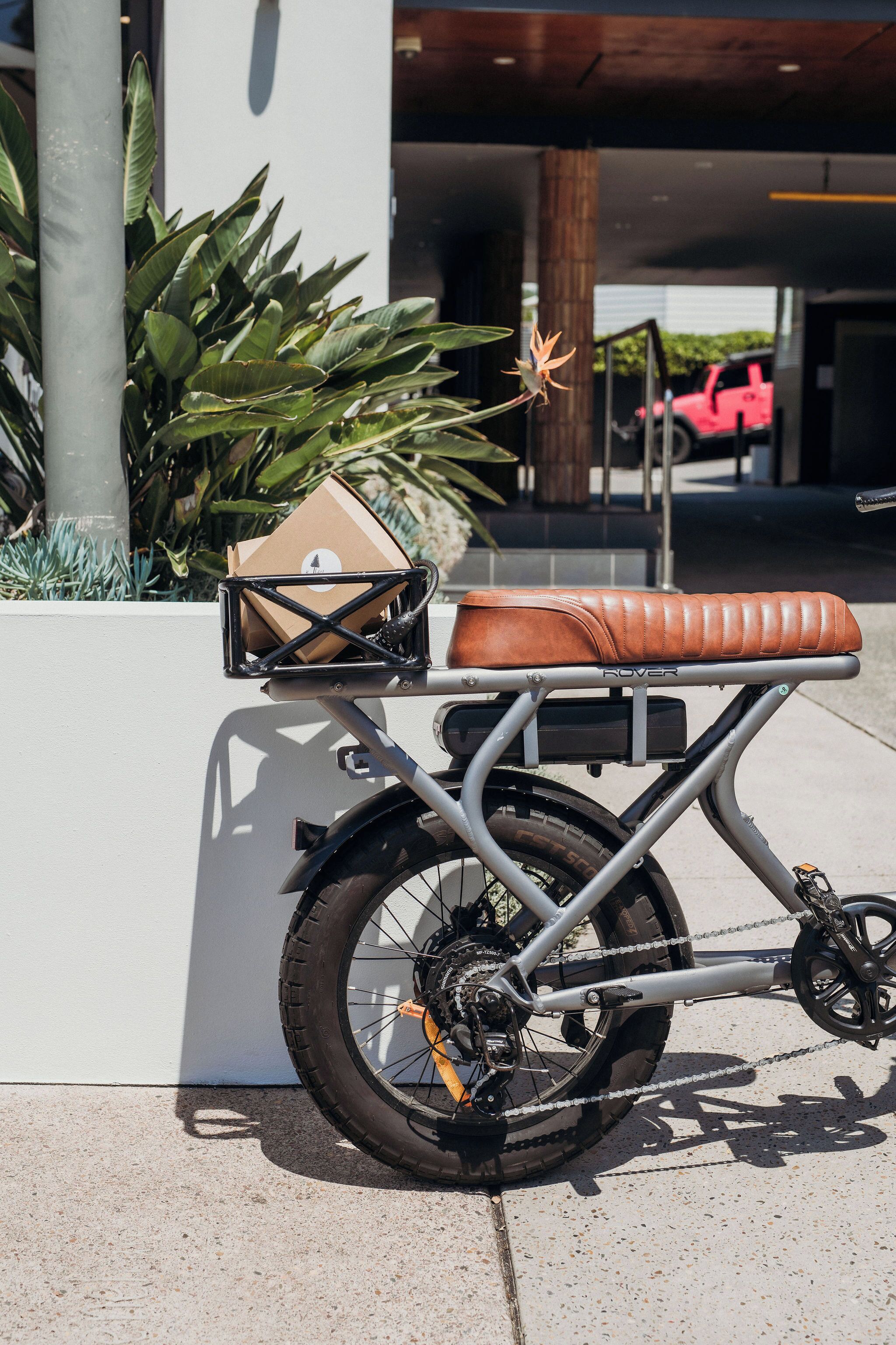 Una bicicleta con asiento de cuero marrón y una caja de cartón en la parrilla trasera, estacionada en una acera junto a un edificio.