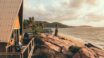 A beachfront property with a modern house, a balcony, and a view of the ocean and hills.