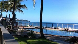 Zona de piscina con sillas de descanso y palmeras, con vista a un mar azul y despejado.