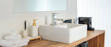 Een moderne badkamer met een houten wastafelmeubel, een witte wastafel en een grote spiegel. Er liggen handdoeken en toiletartikelen op het meubel, en een koffieapparaat is zichtbaar.
