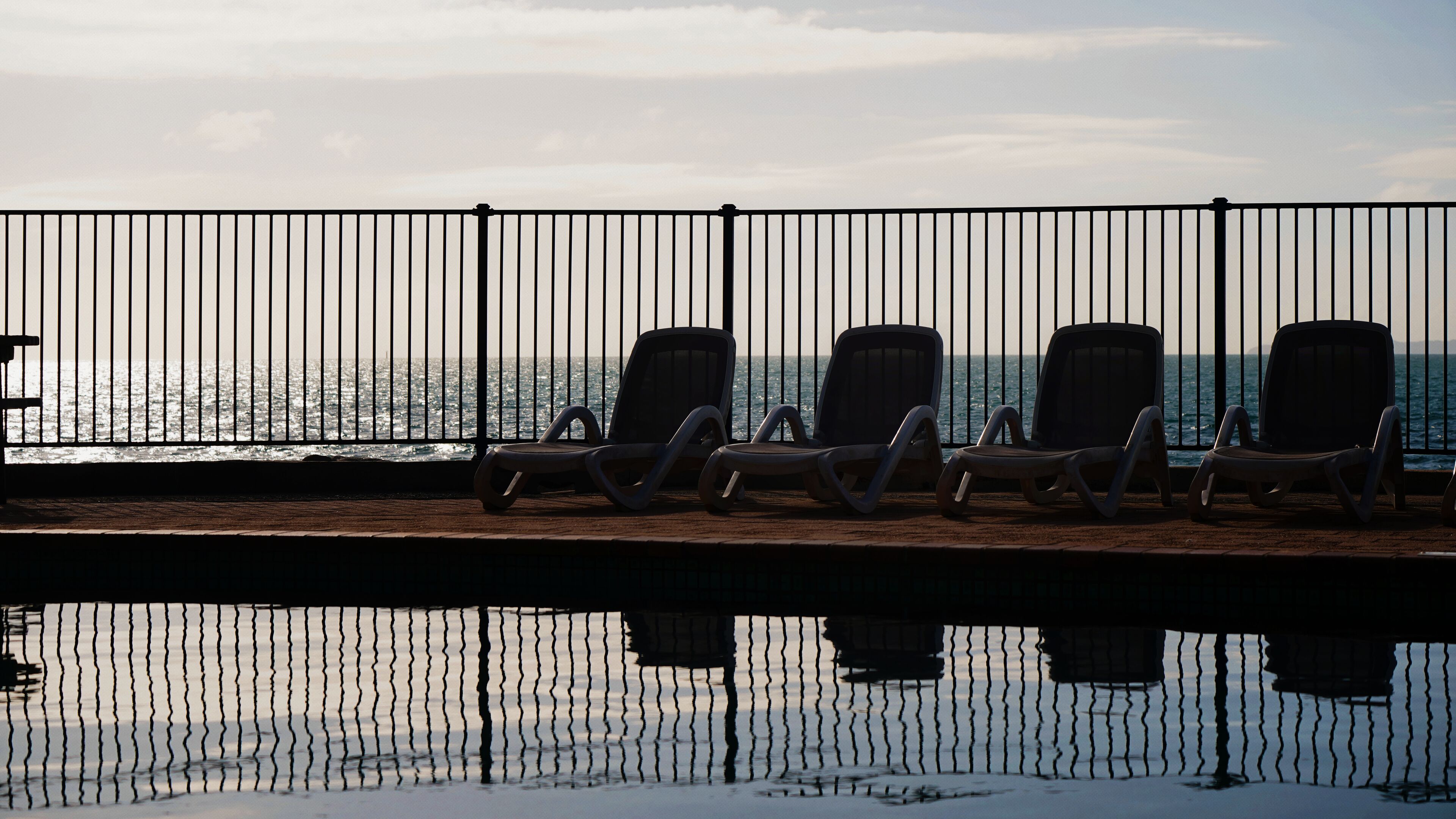 Een rij lege lounge stoelen gericht op een waterlichaam, gescheiden door een metalen hek.