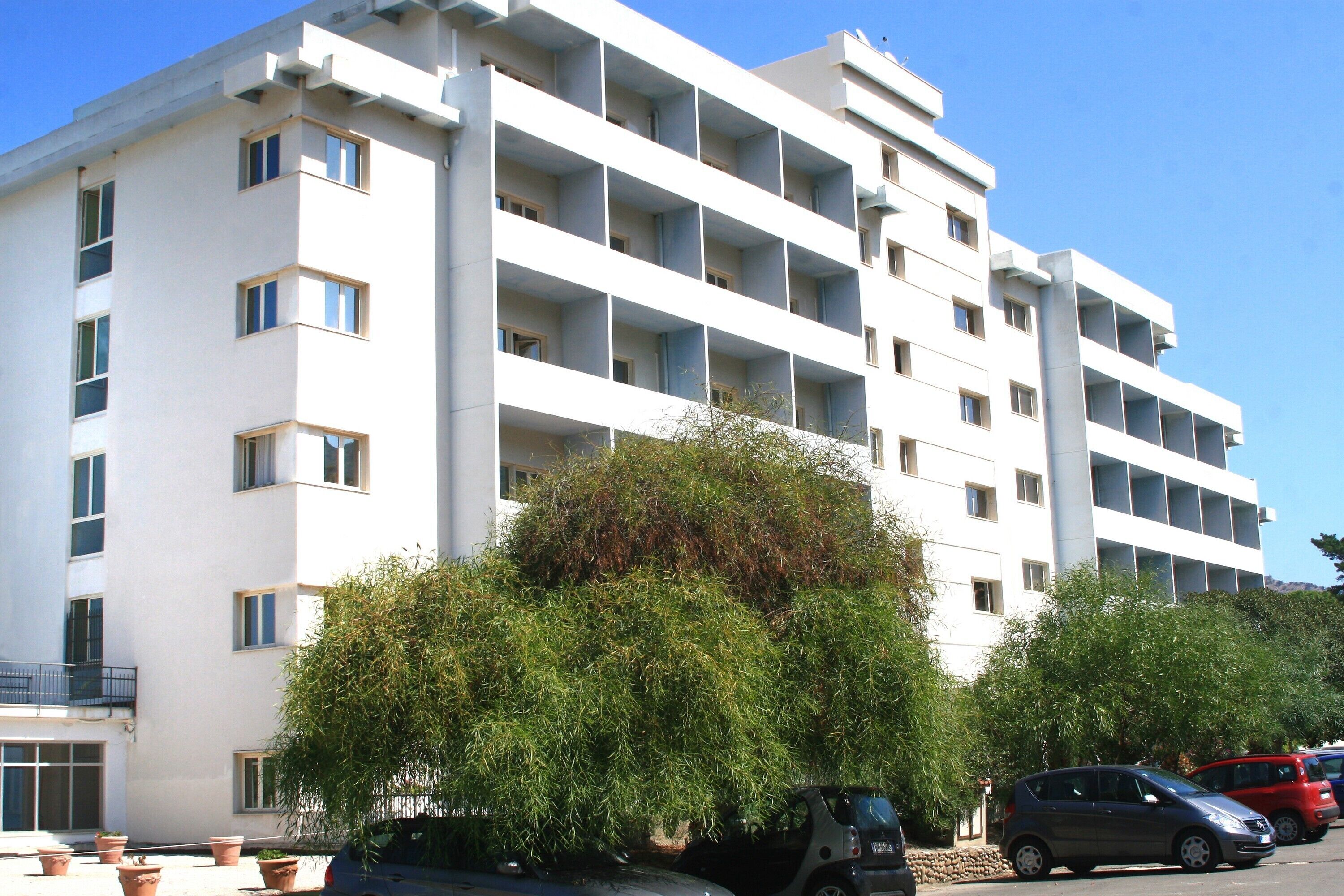 Un edificio de varios pisos con balcones, plantas en macetas y automóviles estacionados.