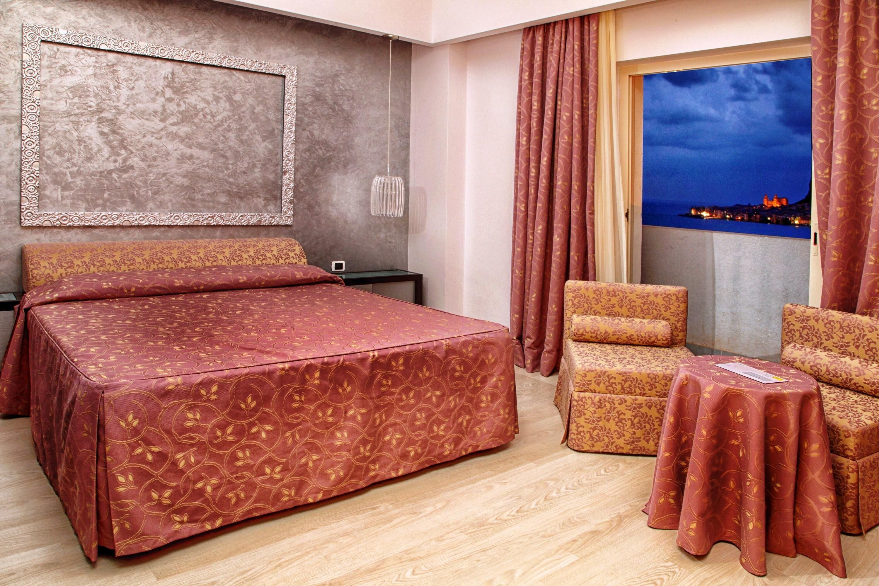 A hotel room with a large bed, two armchairs, a small table, a nightstand, a wall-mounted TV, and a view of a cityscape at dusk.