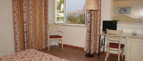 A hotel room with a bed, a desk with a chair, a lamp, and a view of the sea through a window.