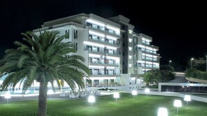 Ein mehrstöckiges Hotel mit modernem Design, das Balkone und einen gut beleuchteten Außenbereich mit Palmen und beleuchteten Tischen bietet.