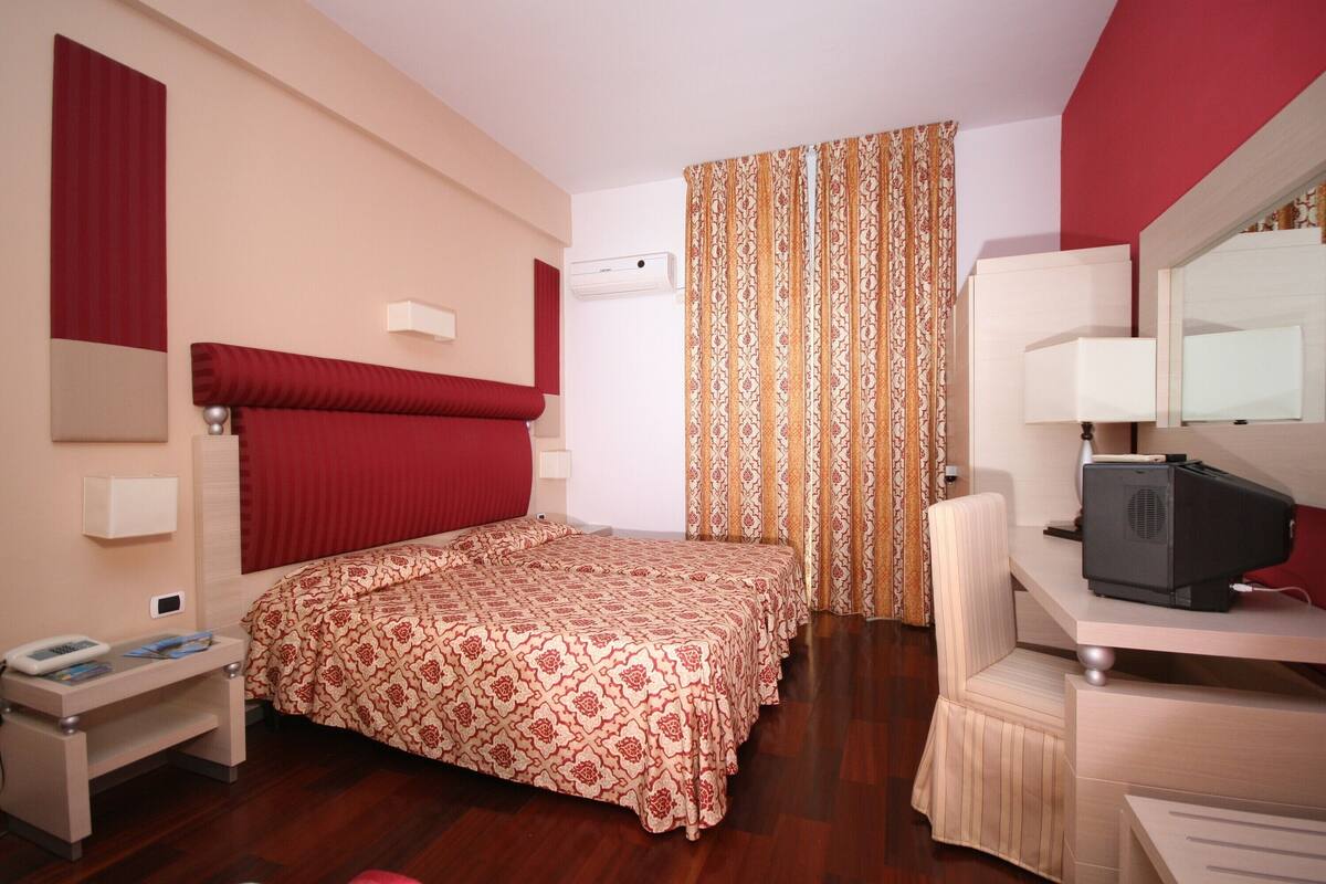 Una habitación de hotel con cama, escritorio con computadora, una silla y ventana con cortinas estampadas.