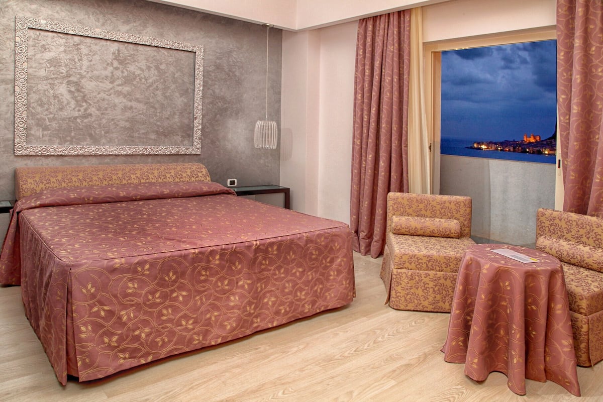 Habitación de hotel con una cama, dos sillones, una mesita, una mesita de noche, un televisor montado en la pared y una ventana con vistas a un paisaje urbano.