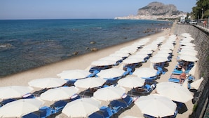 Ein Strand mit Reihen von Sonnenschirmen und Liegestühlen, eine felsige Küstenlinie und eine Stadt im Hintergrund.