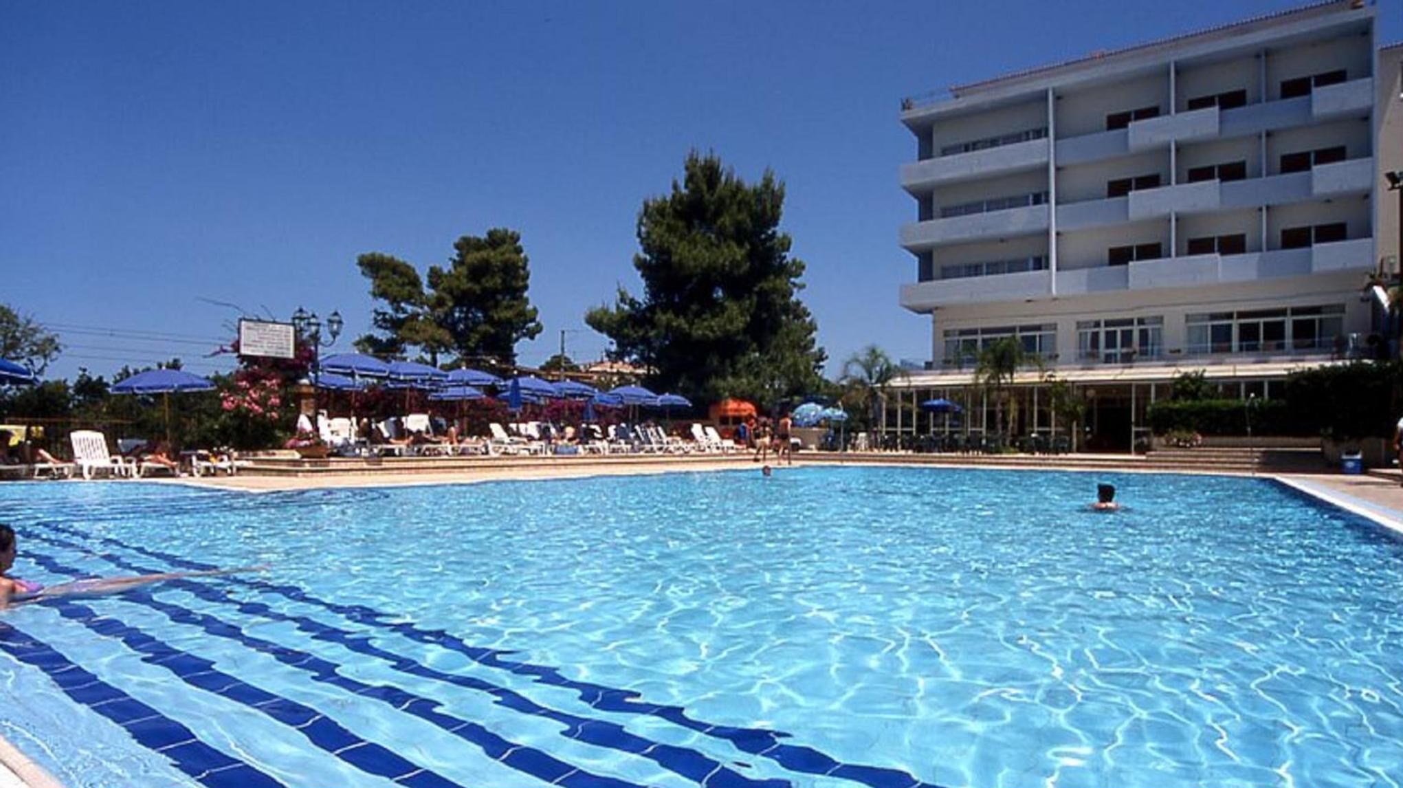 Una piscina con sillones de descanso y sombrillas, rodeada de árboles y un edificio de varios pisos.