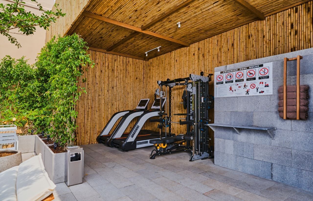 A covered outdoor gym area with exercise equipment, a wall-mounted sign with swimming pool rules, and potted plants.