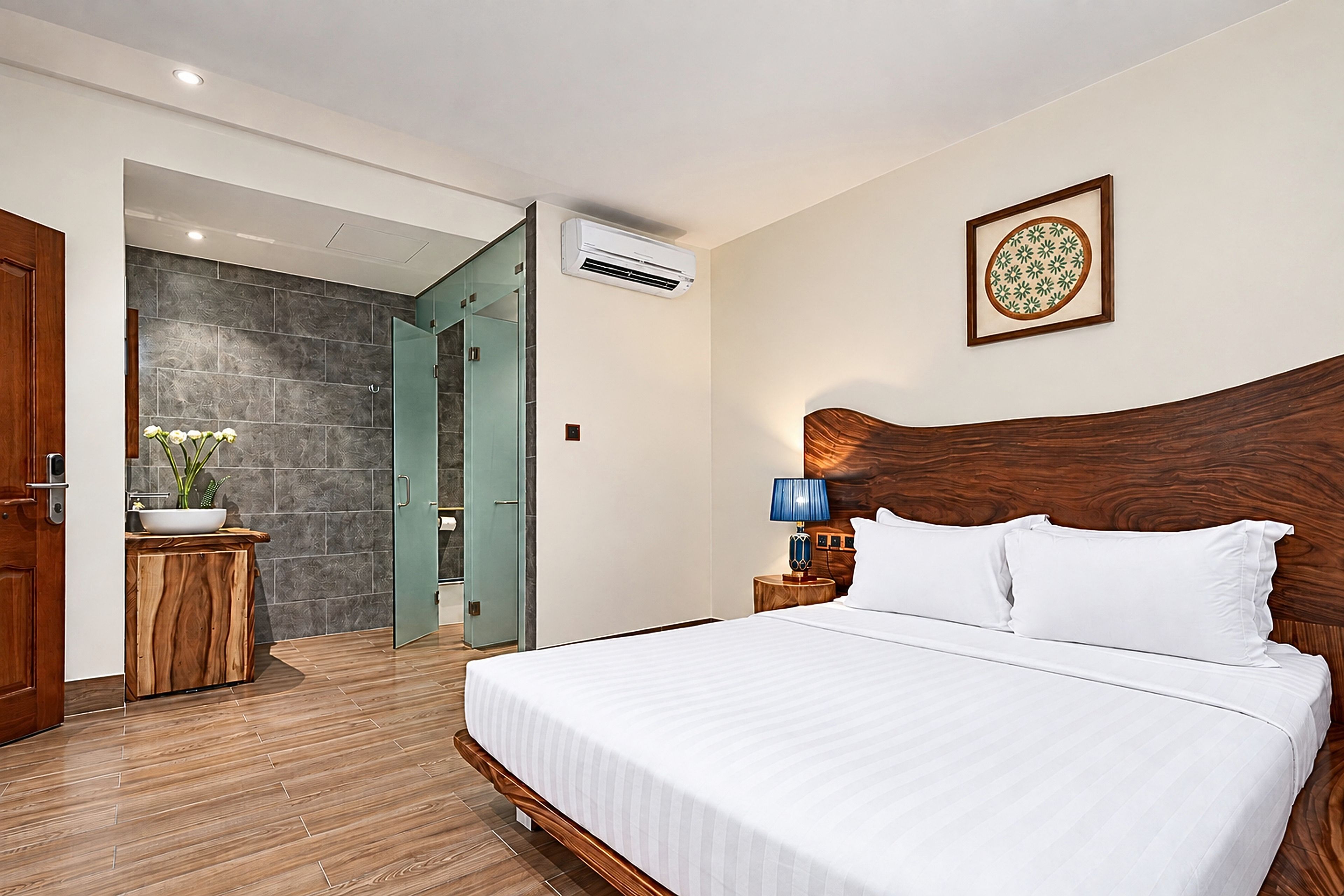 A hotel room with a wooden bed, a nightstand with a lamp, a bathroom with a glass shower enclosure, and a wall-mounted air conditioner.