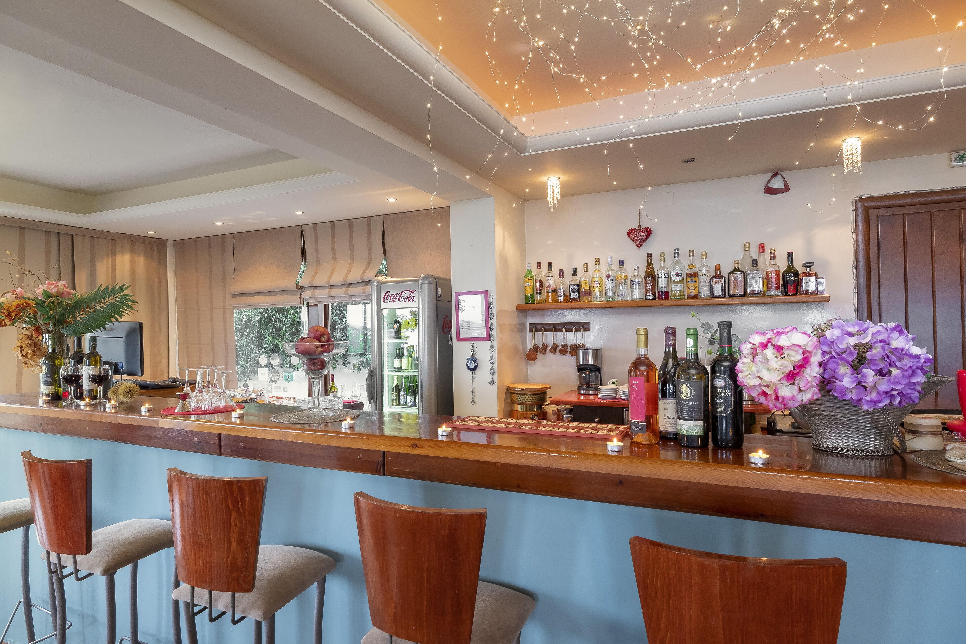 A bar area with wooden bar stools, a refrigerator with a Coca-Cola logo, shelves with various bottles, and a decorative light fixture.