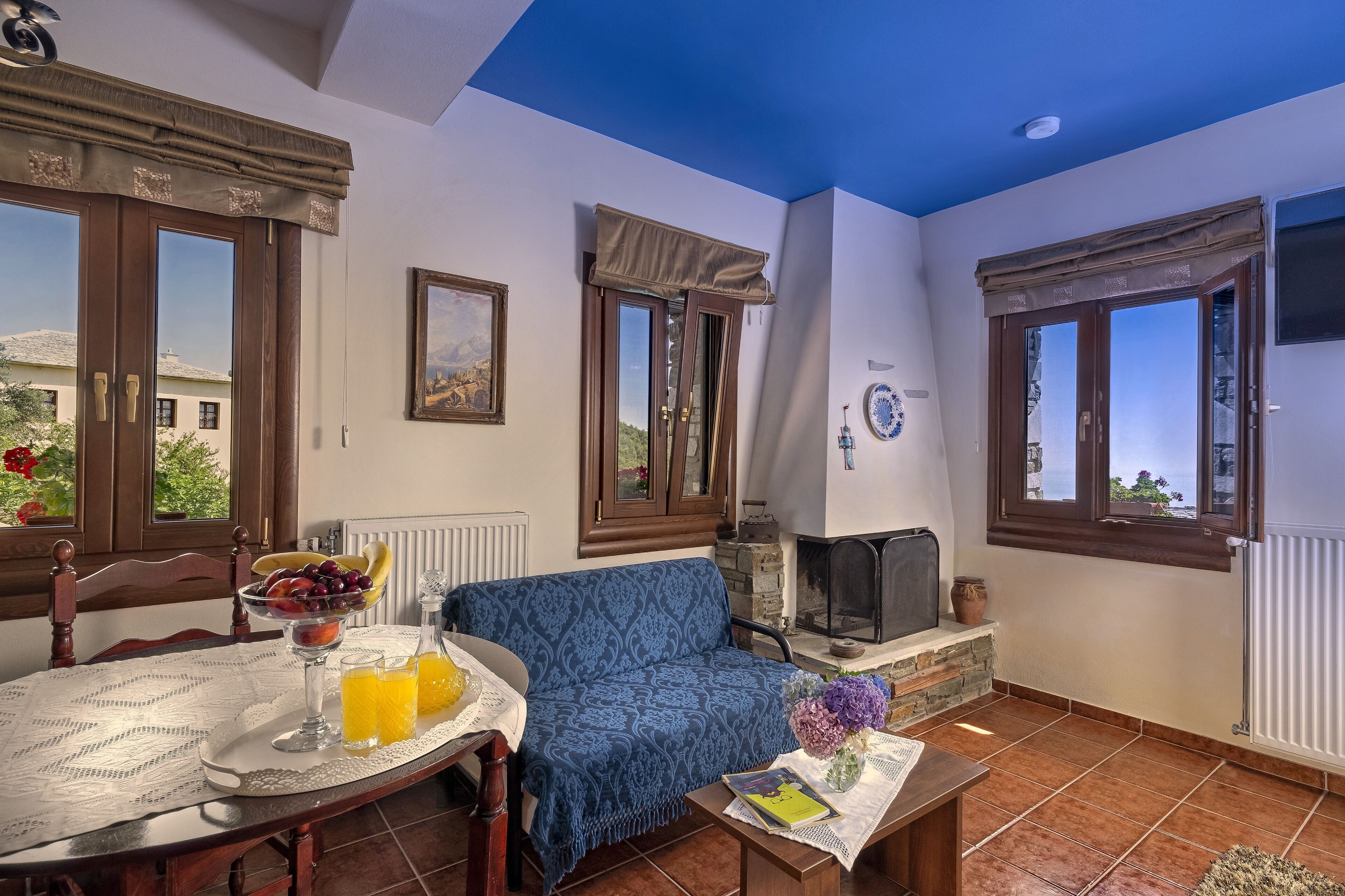 A cozy living room with a blue sofa, a dining table with a fruit bowl and drinks, a fireplace, and large windows with curtains.