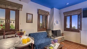A cozy living room with a blue sofa, a dining table with a fruit bowl and drinks, a fireplace, and large windows with curtains.