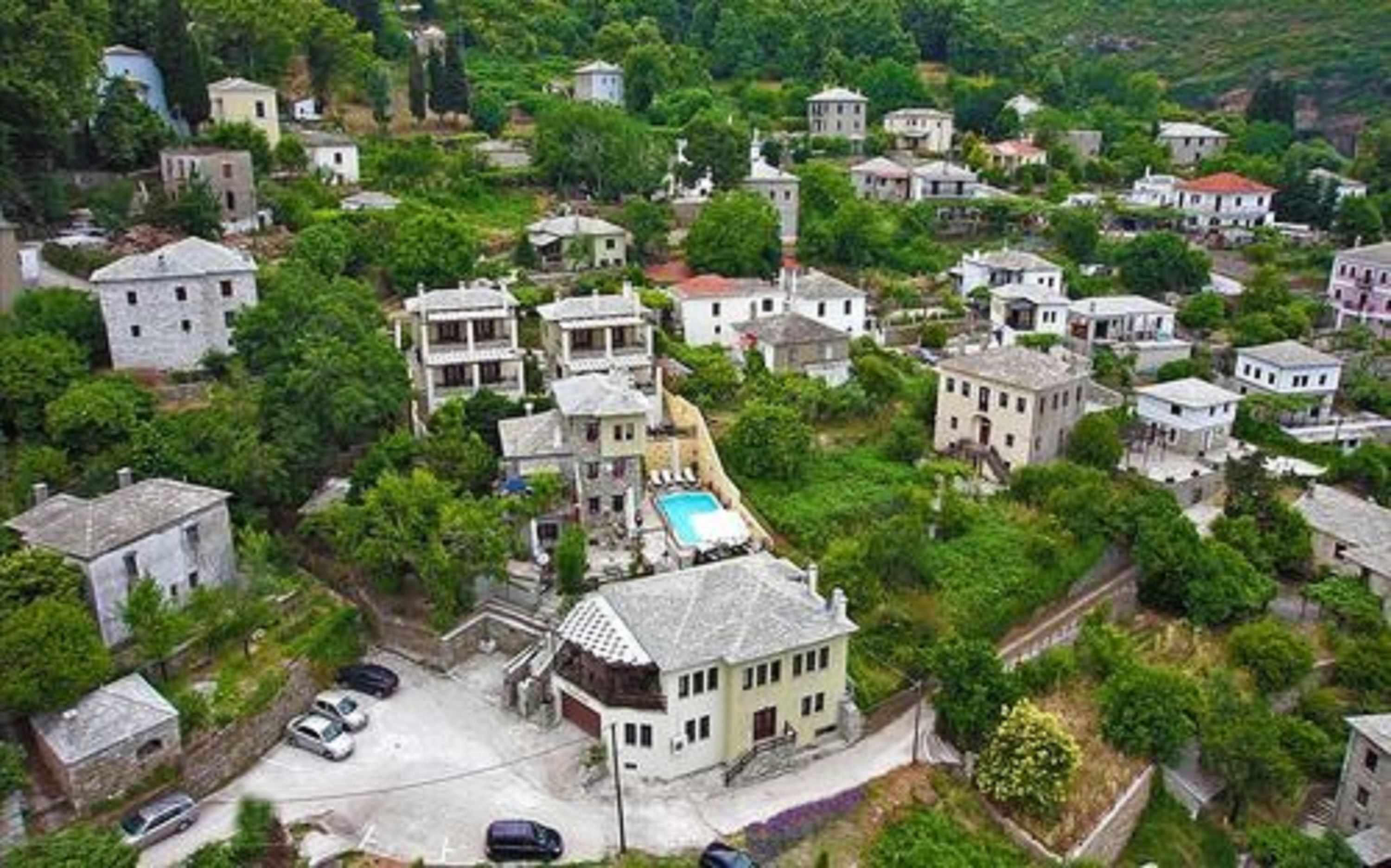 An aerial view of a residential area with various buildings, including a house with a swimming pool, surrounded by greenery.