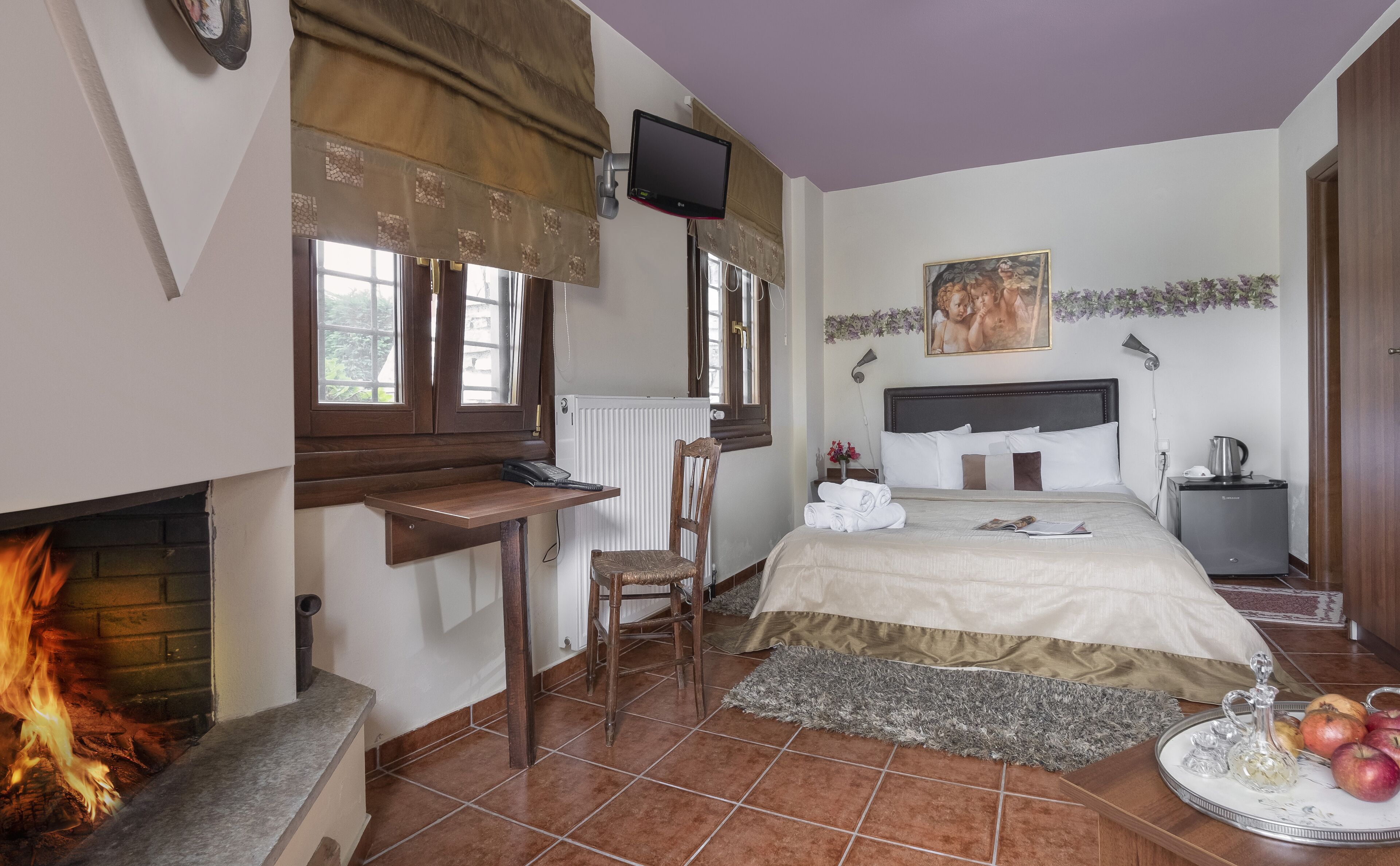 Camera da letto con letto, comodino, sedia, camino, TV e un angolo cottura con bollitore.