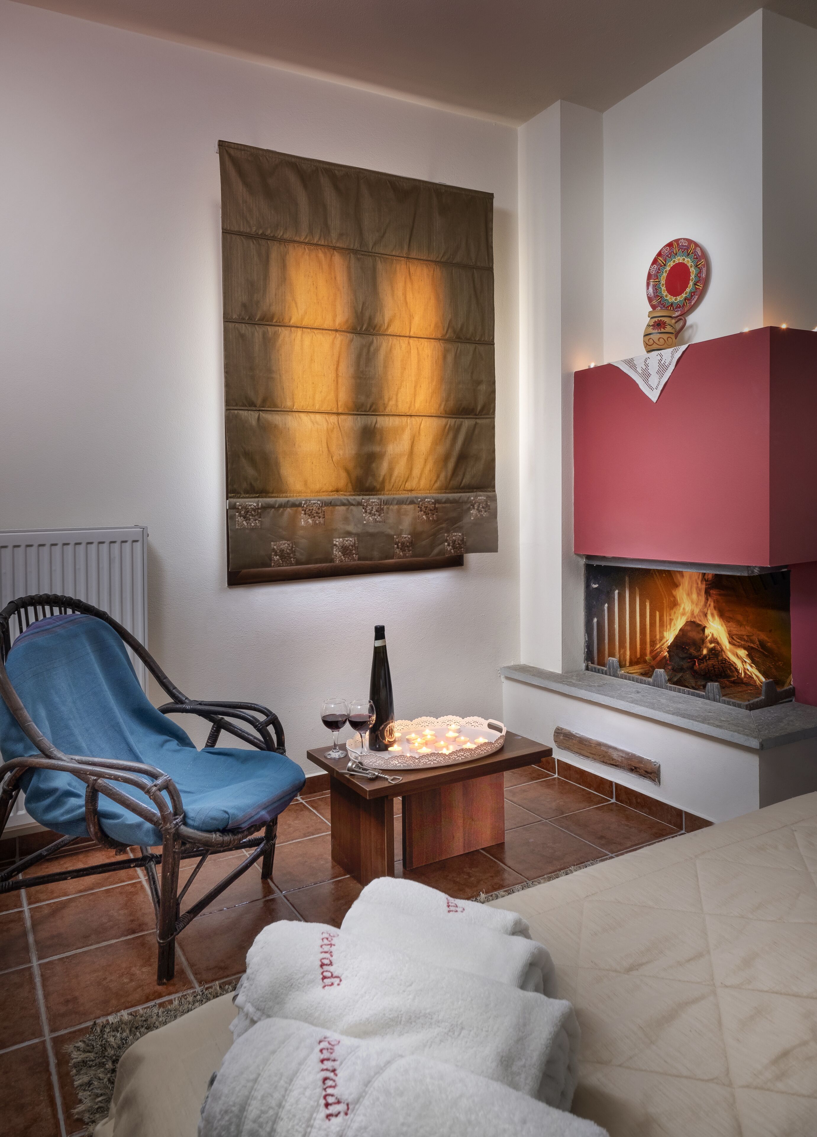 Un salon chaleureux avec une cheminée, un fauteuil bleu, une petite table avec une bouteille et des verres, et une grande fenêtre avec un rideau.