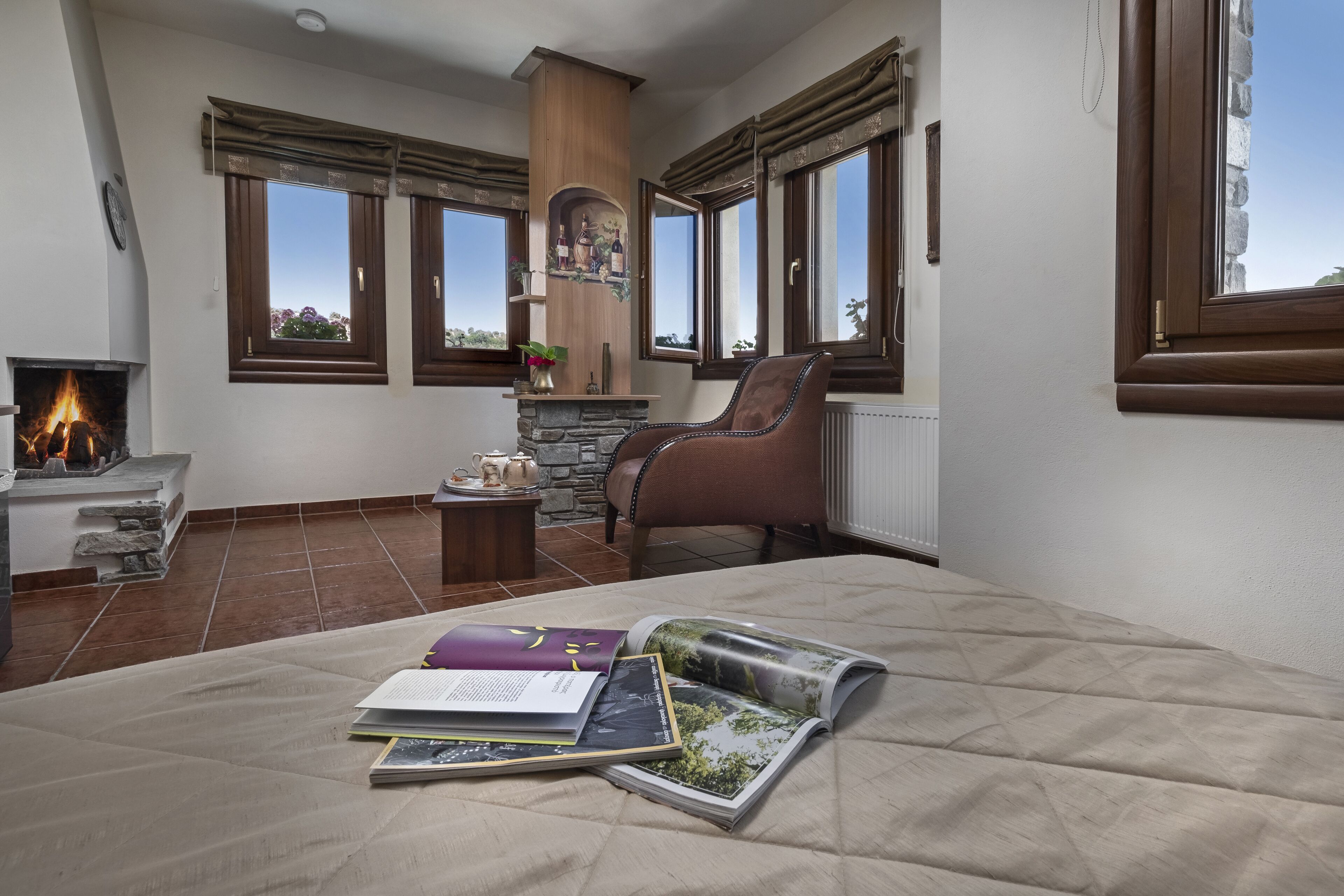 A bedroom with a fireplace, a stone fireplace surround, a large window with a view, a comfortable chair, and a bed with a quilt.