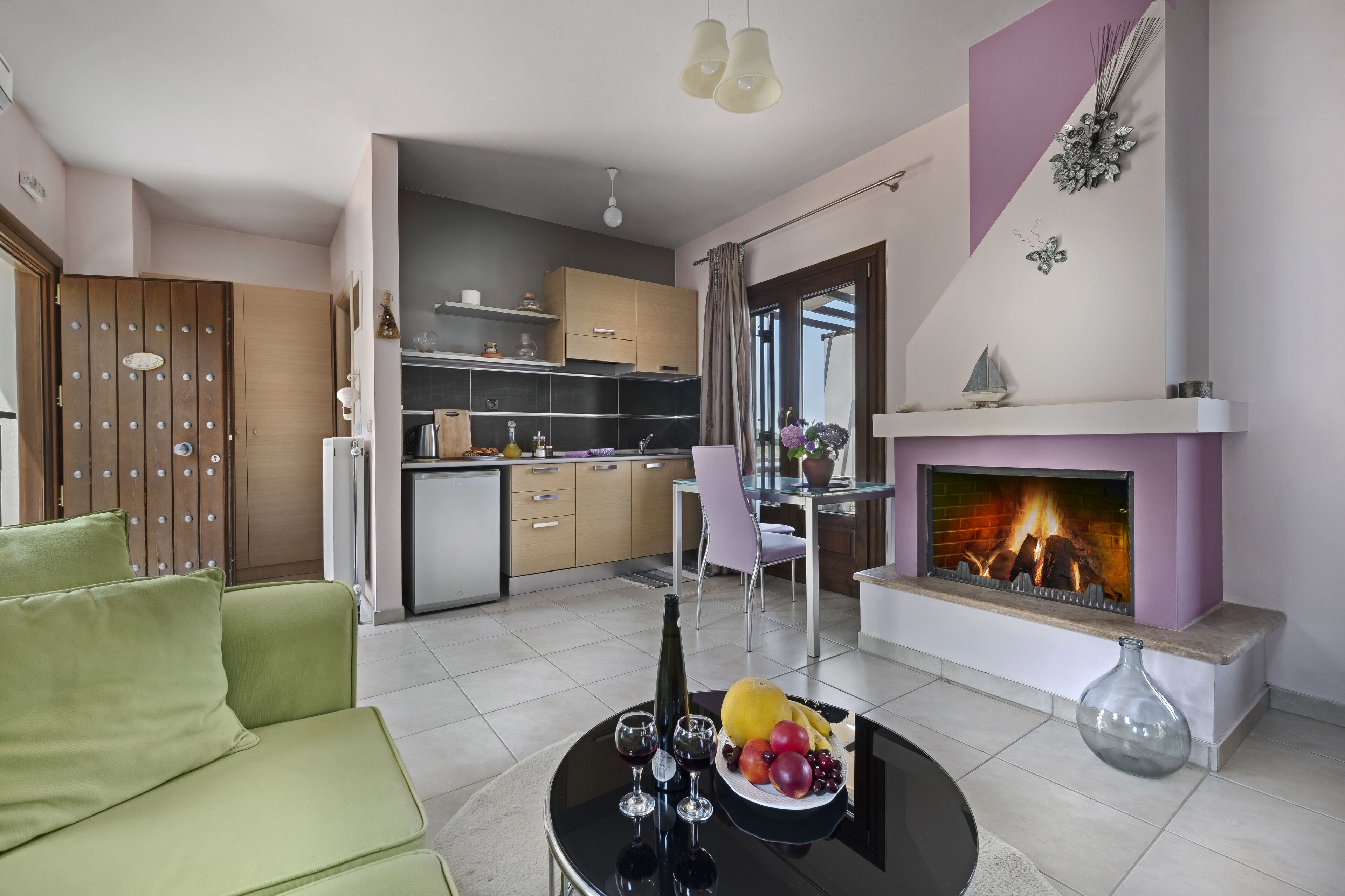 A modern living room with a green sofa, a dining table with a fruit bowl and wine glasses, a fireplace, and a kitchen in the background.