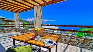 Une table en bois, sur laquelle se trouvent un panier de fruits, une tasse et un journal, est placée sur un balcon offrant une vue panoramique.
