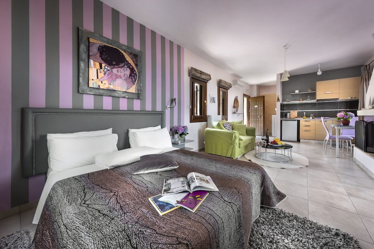 Une chambre d’hôtel moderne avec un grand lit, un fauteuil vert, une petite table à manger et un coin cuisine à l’arrière-plan.