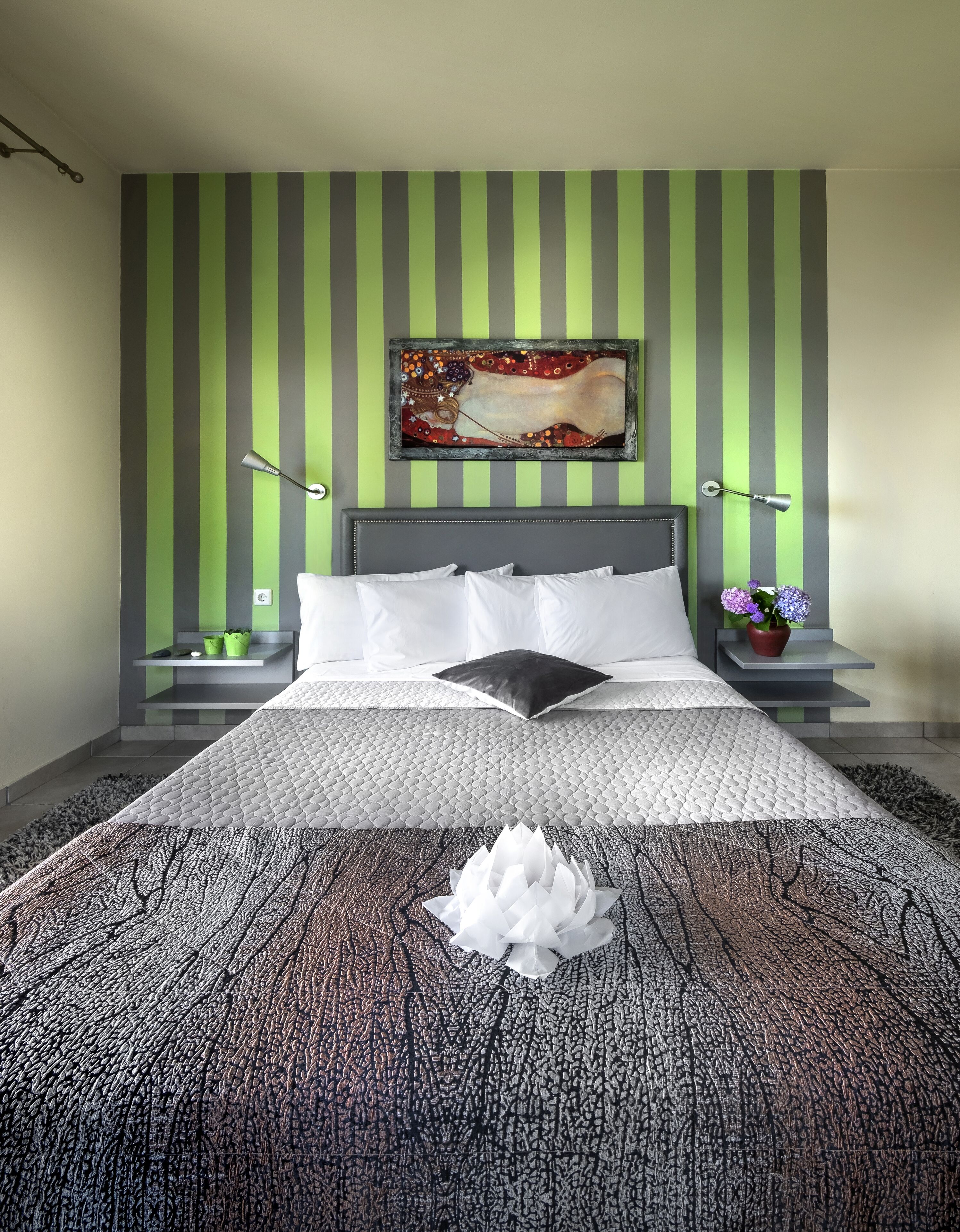 Une chambre moderne avec un grand lit, des murs à rayures vertes et grises, un tableau décoratif, deux tables de chevet avec des lampes et un arrangement floral décoratif.