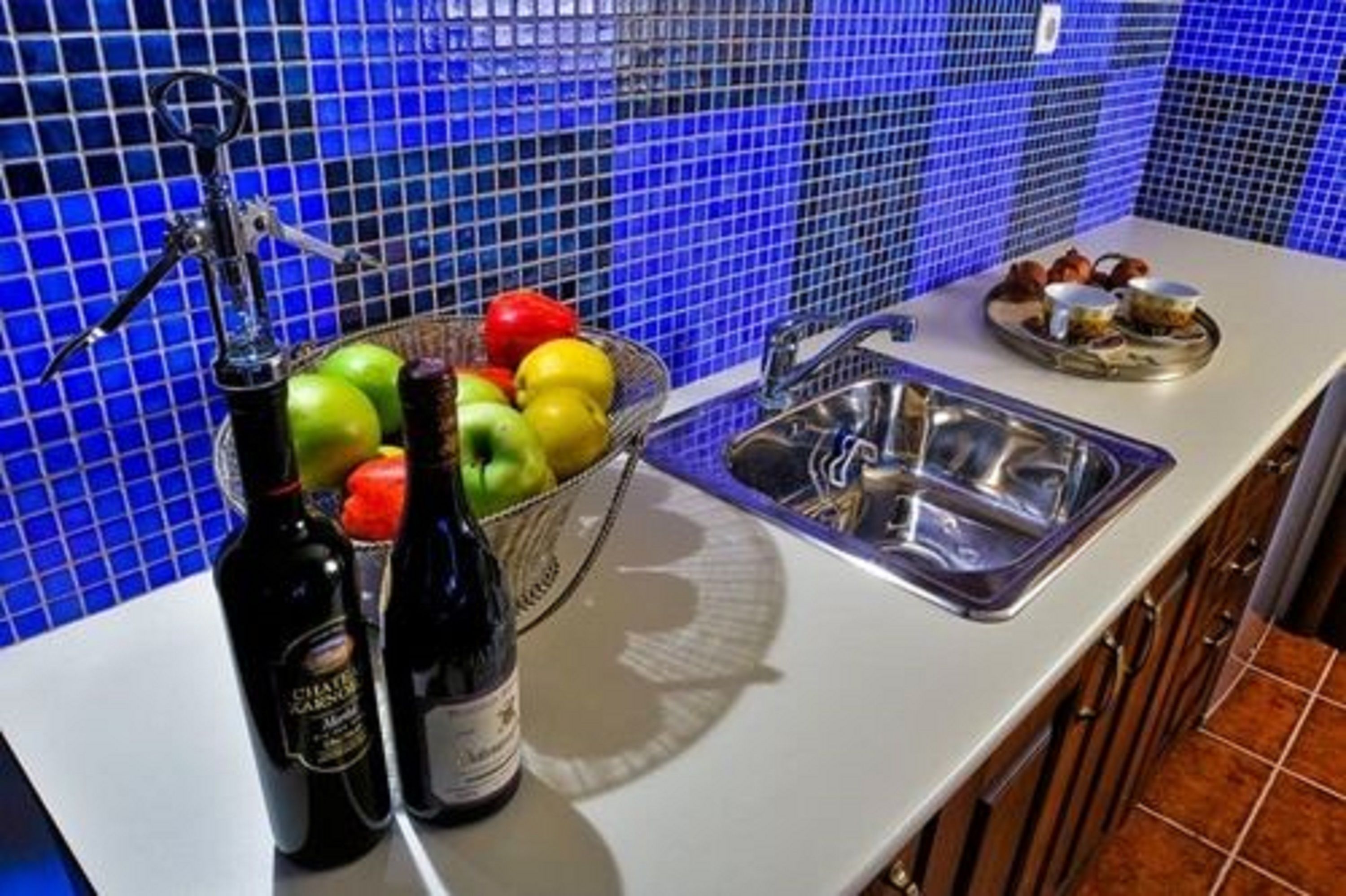 A kitchen with a blue tiled backsplash, a sink, and a fruit bowl containing apples and oranges.