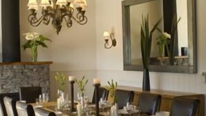 A dining area with a long wooden table, multiple chairs, a chandelier, and a stone fireplace. - The Sanctuary @ Bay of Islands (Opua)