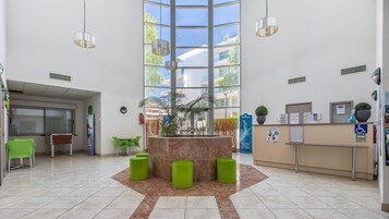 Un hall d’entrée spacieux comprenant un comptoir d’accueil, des sièges verts, un grand jardinier central et de grandes fenêtres offrant une vue sur l’extérieur.