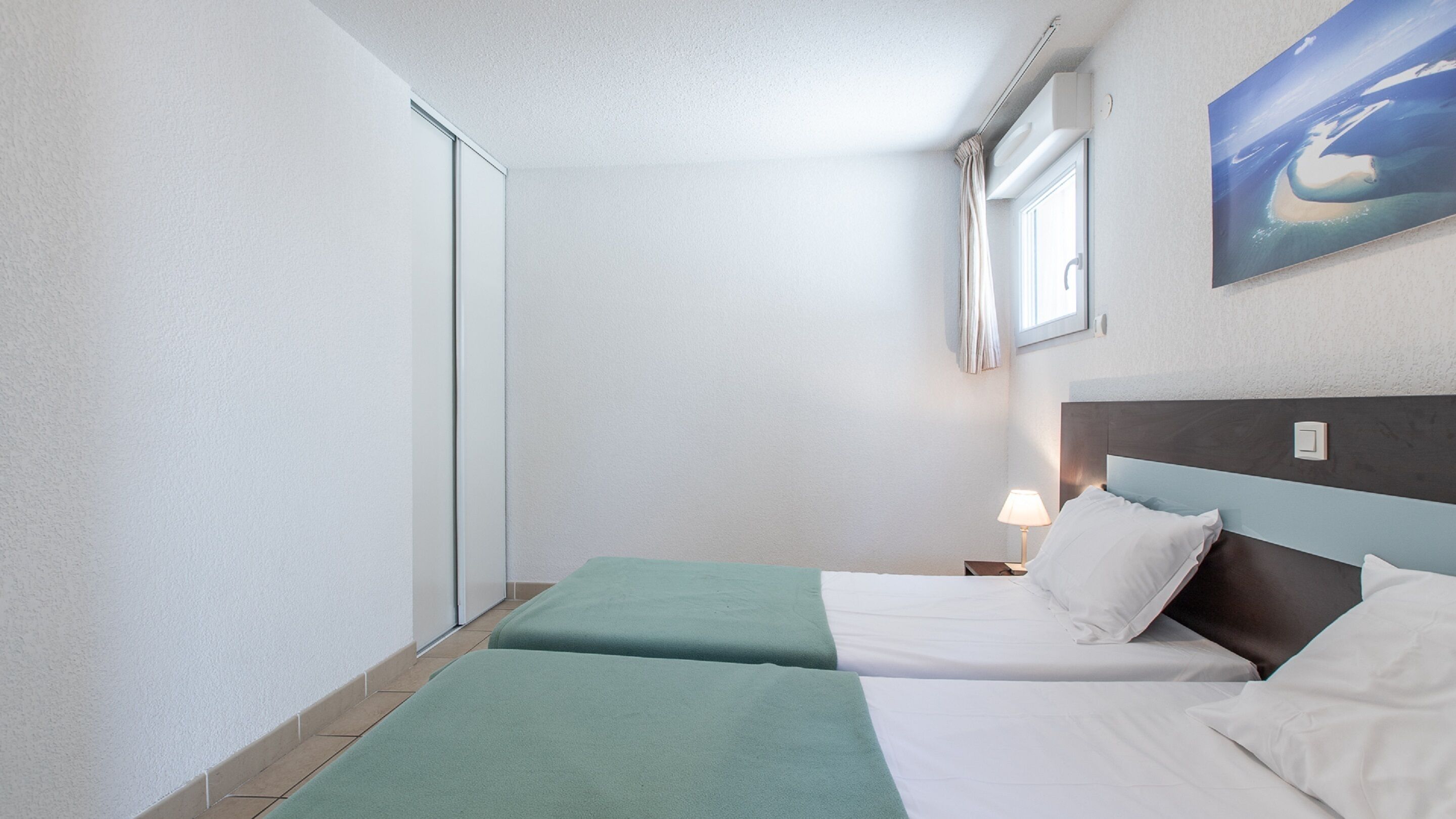 Une chambre d’hôtel avec deux lits, une fenêtre avec des rideaux, un tableau au mur et une table de chevet avec une lampe.