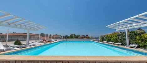 A rectangular swimming pool with white sun loungers and a pergola on one side.