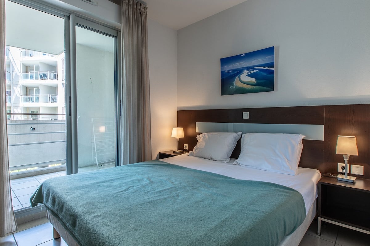 Une chambre d’hôtel avec un lit, deux tables de chevet avec des lampes, un tableau au mur et une vue sur un immeuble par une porte coulissante.