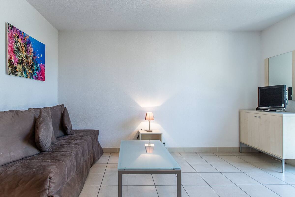 Un salon moderne avec un canapé marron, une table basse en verre, une lampe, un téléviseur posé sur un meuble et un tableau coloré accroché au mur.