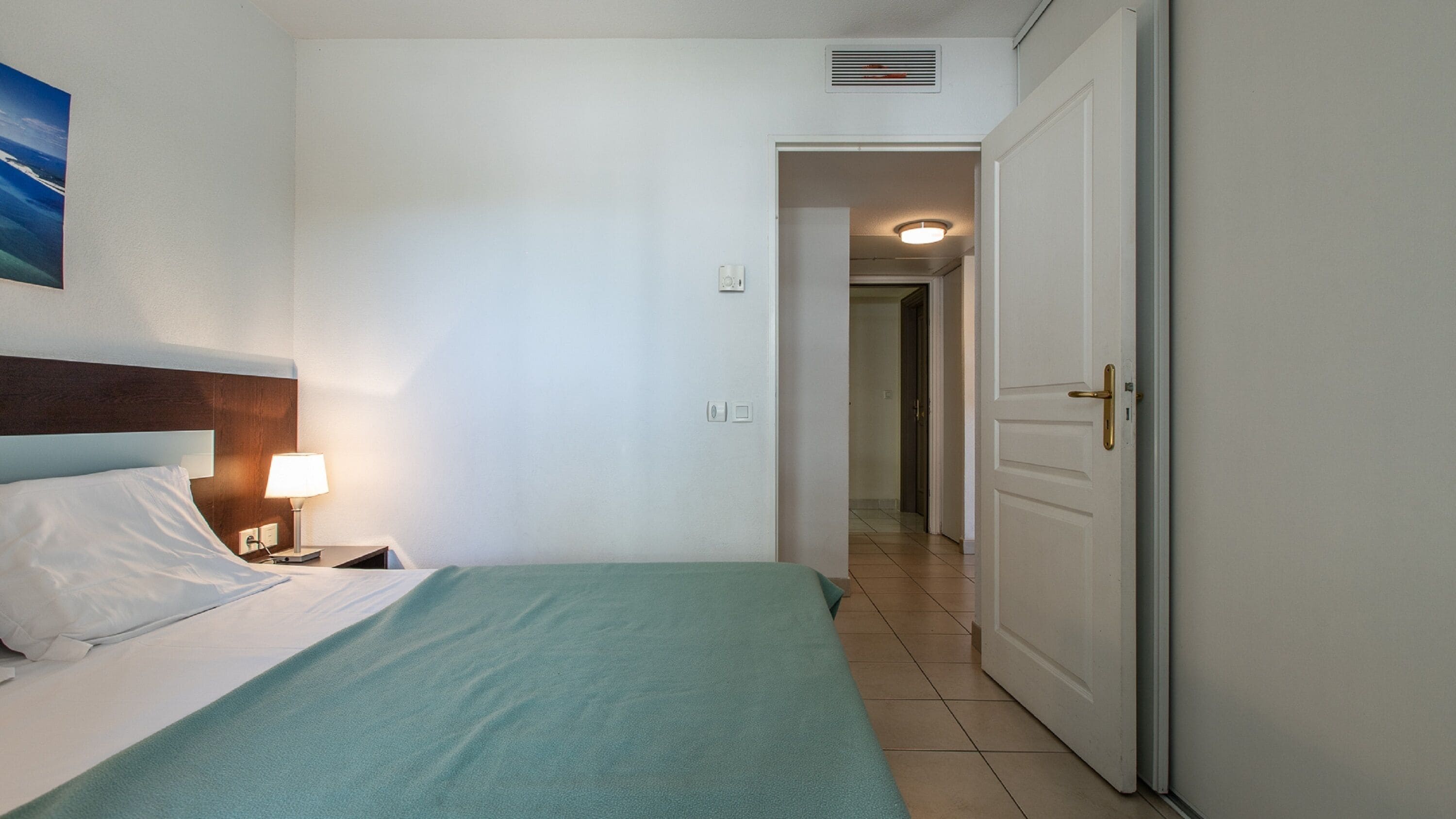 Una habitación de hotel con cama, mesita de noche, lámpara, puerta y pasillo que conduce a otra habitación.