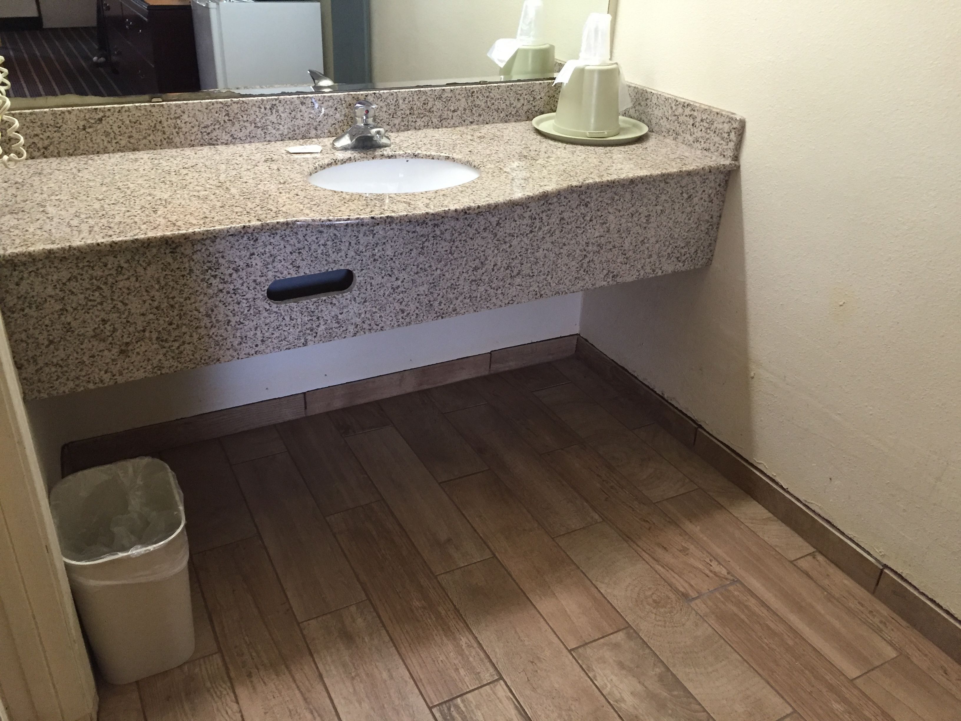 Un baño con encimera de granito, un lavamanos, un espejo, dispensador de toallas, un cesto de basura y suelo de madera.