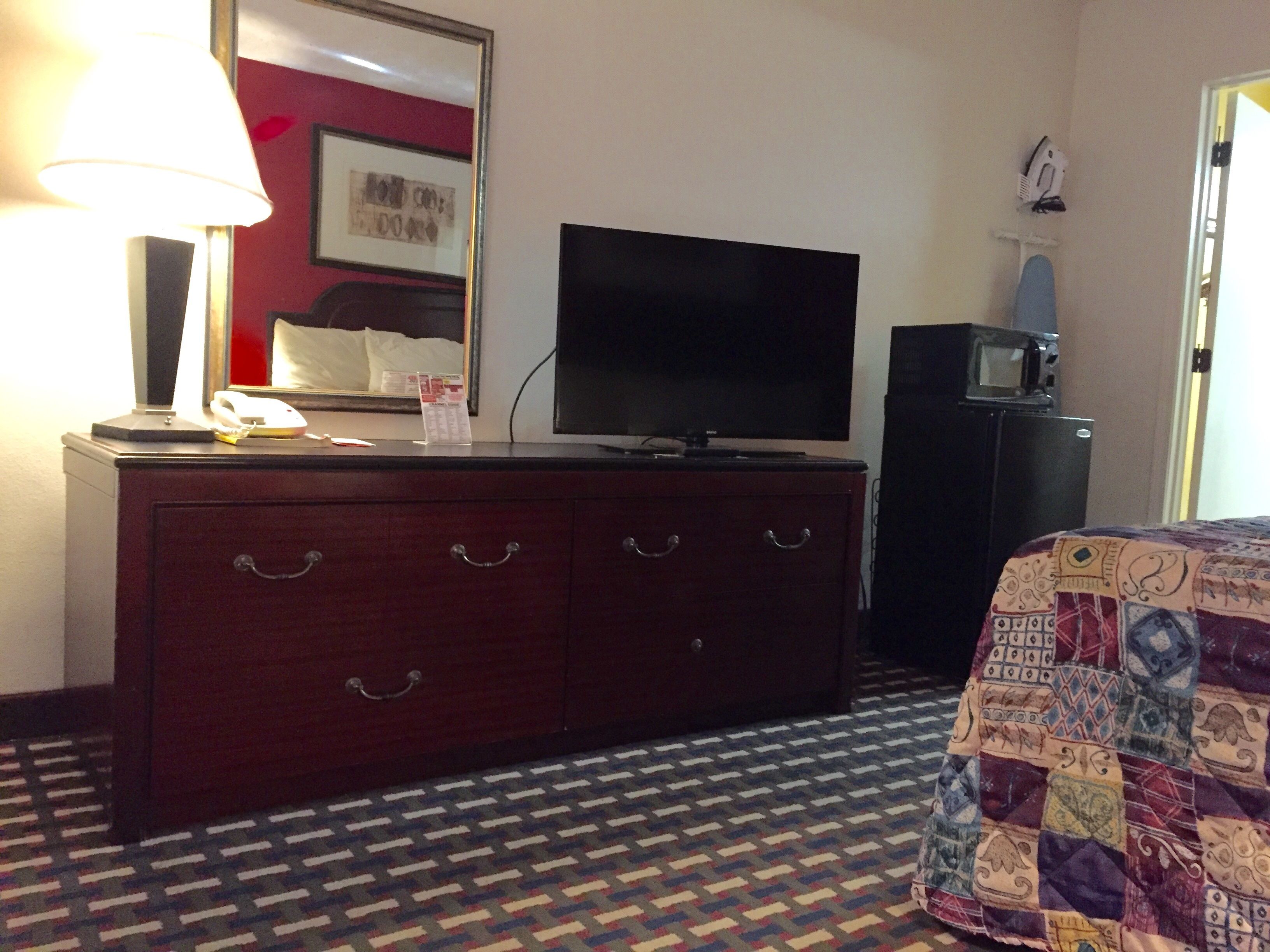 A hotel room with a wooden dresser, a flat-screen TV, a microwave, and a patterned carpet.