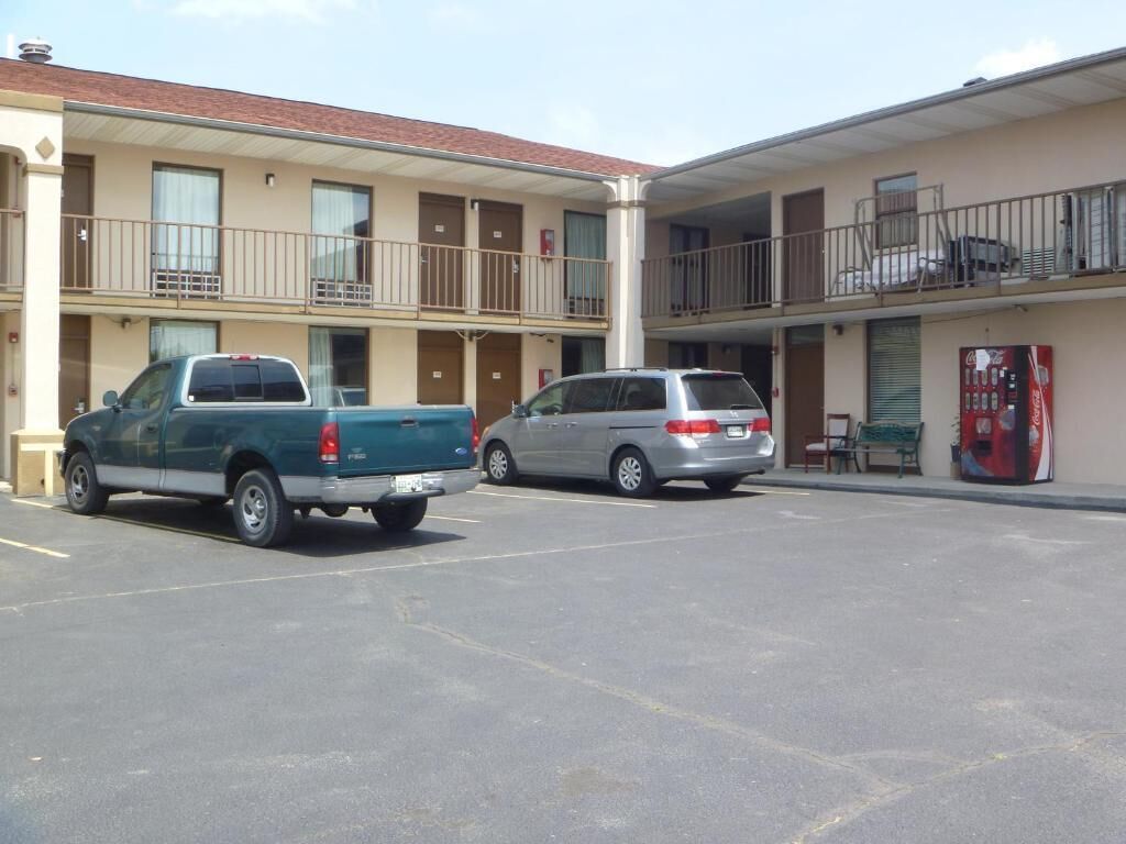 A parking lot with a green pickup truck and a silver minivan parked in front of a motel with a balcony on the second floor.