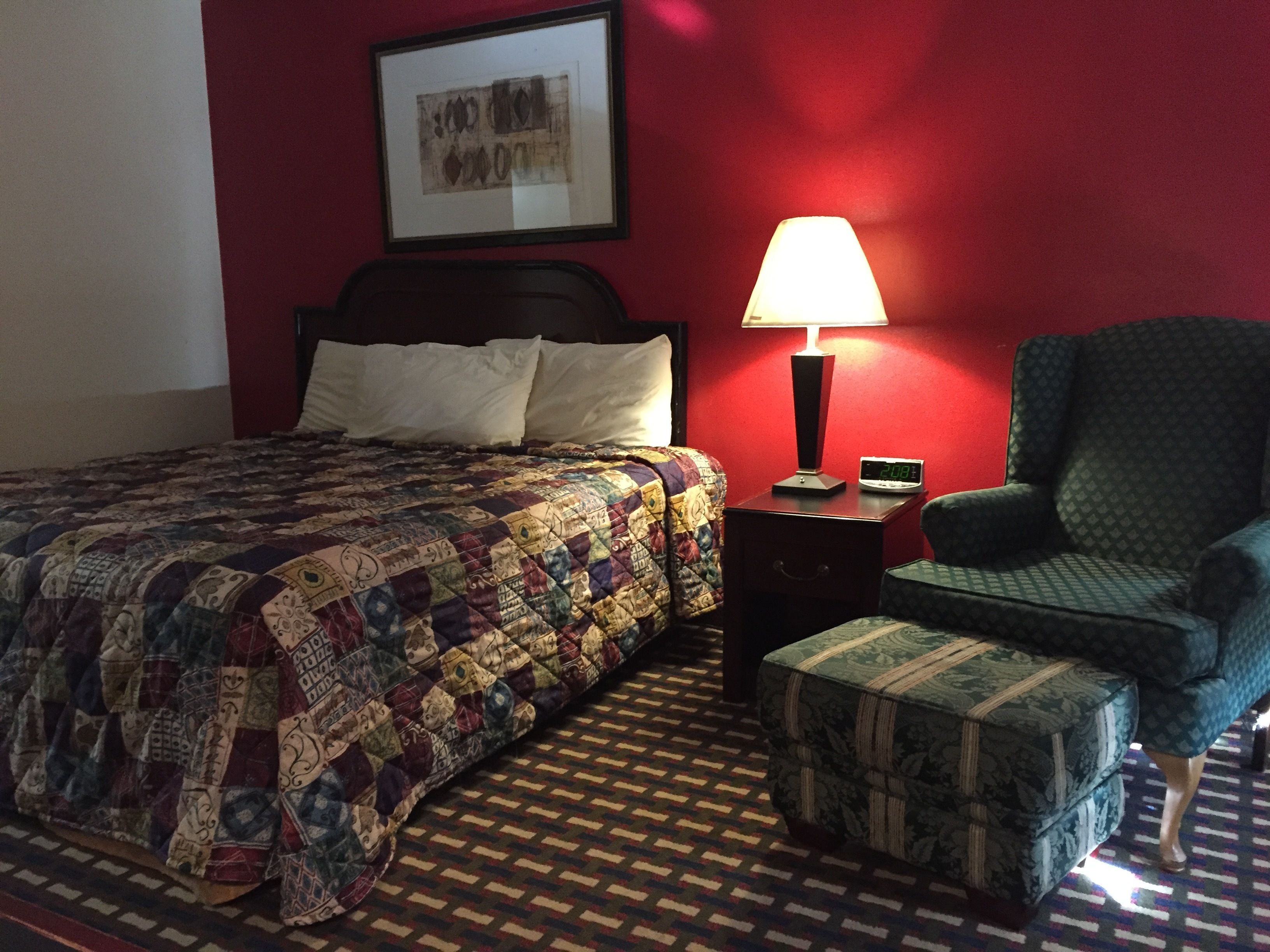 Habitación de hotel con cama, mesita de noche con lámpara, una silla y una alfombra estampada.
