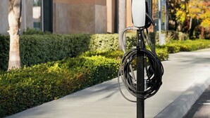 Una estación de carga para vehículos eléctricos en una acera, con un edificio y follaje al fondo.