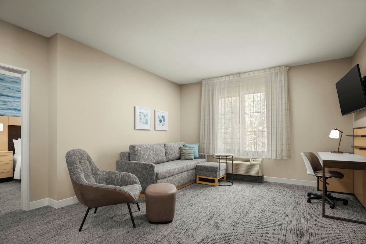 A hotel room with a grey sofa, two armchairs, a small ottoman, a desk with a chair, a flat-screen TV, and a window with sheer curtains.