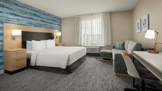 Habitación de hotel con cama, mesitas de noche, escritorio, silla, sofá, ventana con cortinas y obras de arte en la pared.