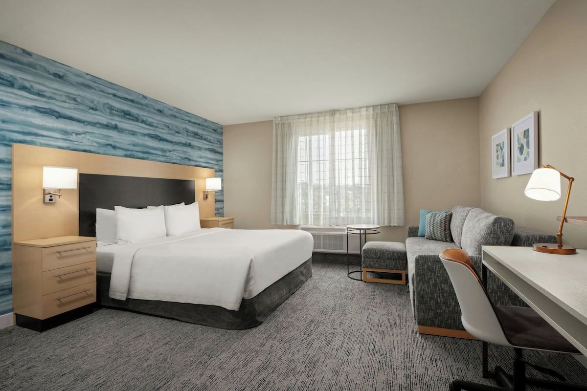 A hotel room with a bed, bedside tables, a desk, a chair, a sofa, a window with curtains, and artwork on the wall.